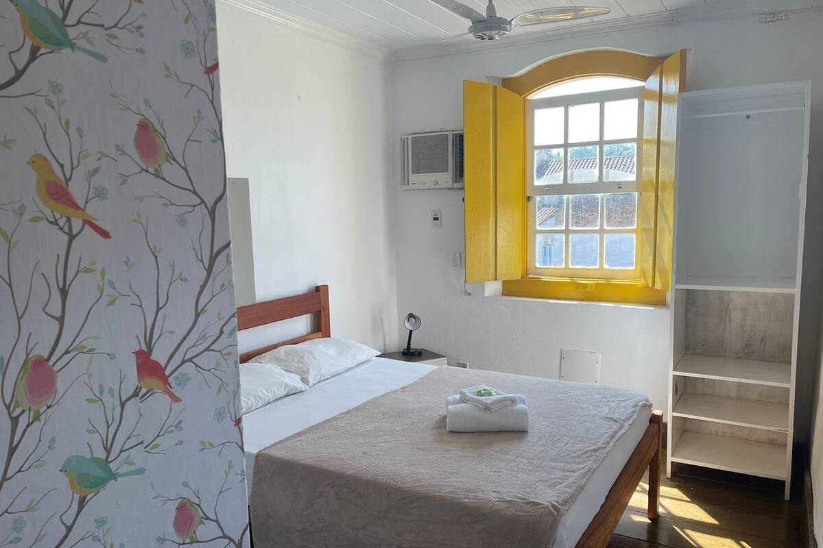 Ein Schlafzimmer mit einem Bett, einem Fenster mit gelben Fensterläden, einem Deckenventilator und einem Kleiderschrank.