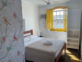 Un dormitorio con cama, una ventana con contraventanas amarillas, un ventilador de techo y un armario.