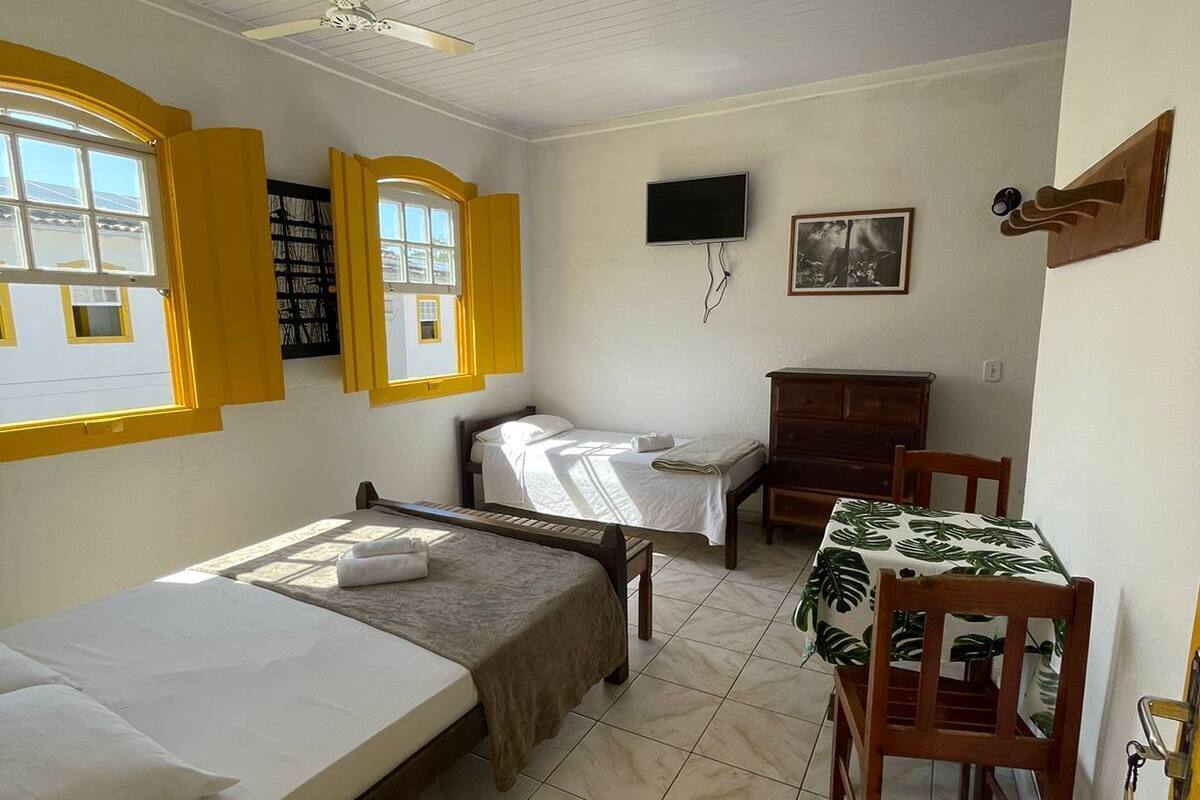 Ein Zimmer mit zwei Einzelbetten, einem Schreibtisch mit Stuhl, einem Fernseher und einem Fenster mit gelben Fensterläden.