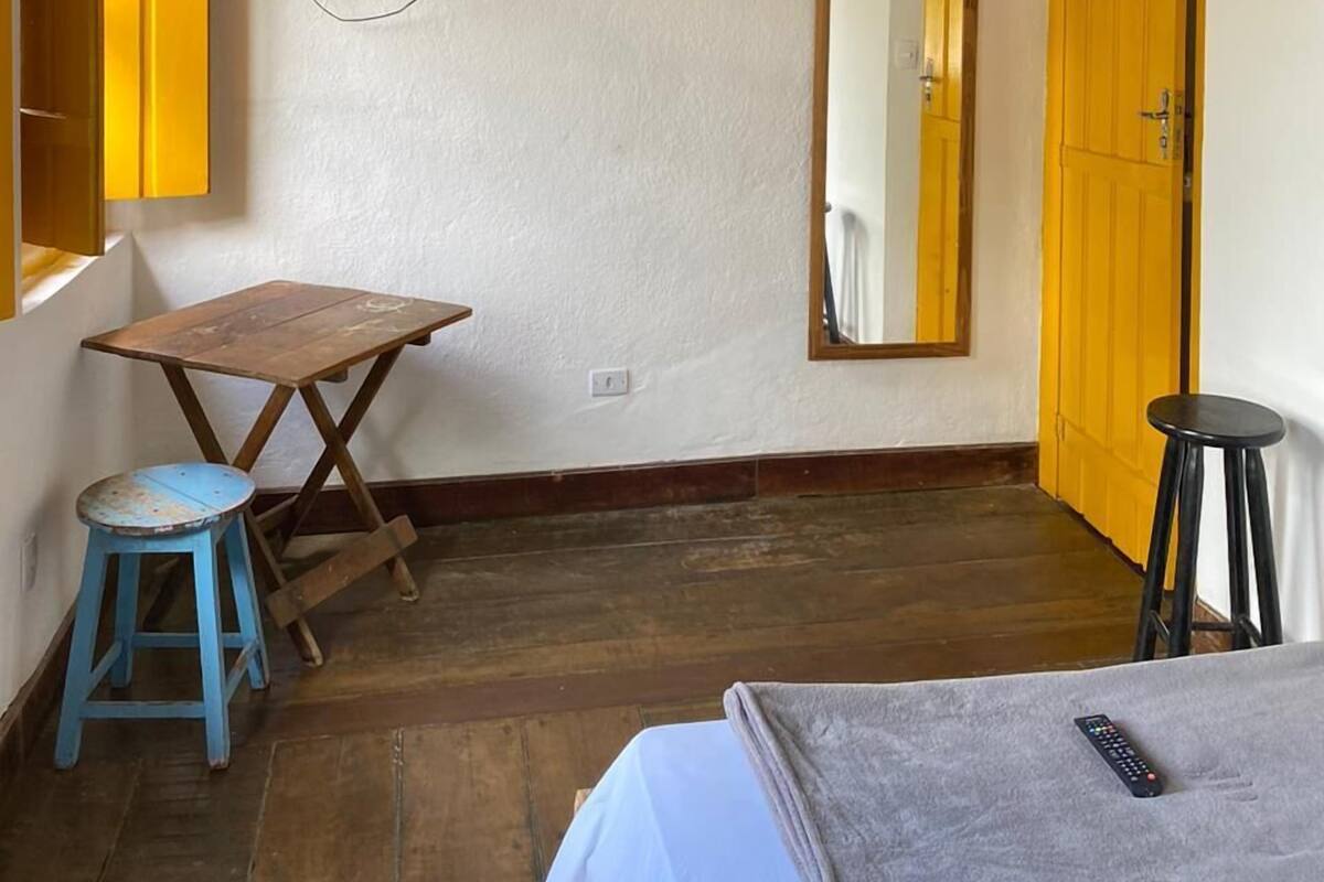 Ein Zimmer mit Holzboden, einem an der Wand befestigten Fernseher, einem Spiegel, einer gelben Tür, einem kleinen Tisch mit Hocker und einem Bett mit Decke.