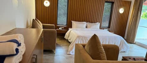 Une chambre d’hôtel moderne avec un mur lambrissé en bois, un grand lit avec du linge de maison blanc, un canapé, une petite table avec des serviettes et une fenêtre avec des stores.