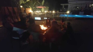 Un grupo de personas reunidas alrededor de un brasero en un entorno al aire libre, con una piscina y un edificio al fondo.