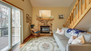 A cozy living room with a stone fireplace, a wooden staircase, and a large window overlooking a deck. - Charming Family Cabin in Crosslake (Crosslake)