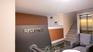 A reception area with a sign, a wooden panel wall, a marble floor, a grey chair, and a staircase with an elevator. - Lara Street Hotel (Antalya)
