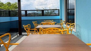 A dining area with a wooden table and yellow chairs, a blue wall, and a view of greenery and buildings. - Hotel Mais Lazer (Joinville)