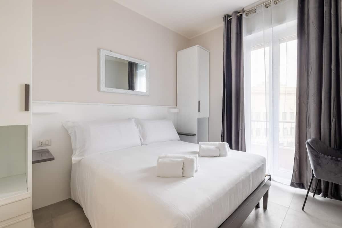 A modern hotel room with a large bed, a bedside table, a wardrobe, a mirror, and a view of the outside through a window with curtains.