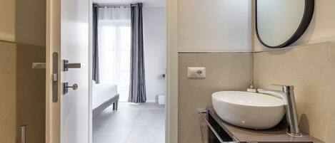 A modern bathroom with a large white sink, a mirror, and a shower area visible through an open door.