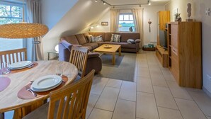 A cozy living room with a sofa, a dining table set for two, and a television on a wooden cabinet. - Vacation apartment Deichblick with sauna (Norddeich)