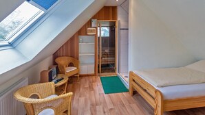 A small, cozy room with a sloped ceiling, a bed, a wicker chair, a small table, and a shower area. - Vacation apartment Deichblick with sauna (Norddeich)