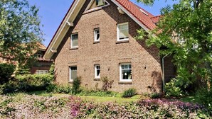 A two-story brick building with a red roof, surrounded by lush greenery and flowering plants. - Vacation apartment Sea Breeze (Norden)