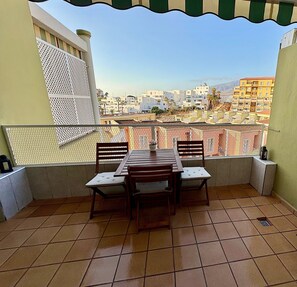 Un balcón con mesa y sillas, vista a un paisaje urbano y una barandilla de seguridad de malla.