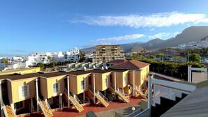 Un edificio costero con varios balcones y un techo rojo, rodeado de edificios blancos y montañas al fondo.