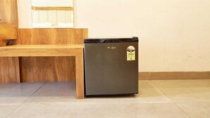 Ein kompakter, schwarzer Kühlschrank mit Aufkleber an der Vorderseite, steht auf einem Fliesenboden neben einer hölzernen Bank und einer Wand mit einem Fenster.