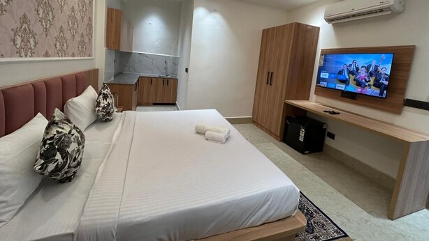 Ein modernes Hotelzimmer mit einem großen Bett, einem Fernseher, einem Holzschrank und einer Küchenzeile im Hintergrund.