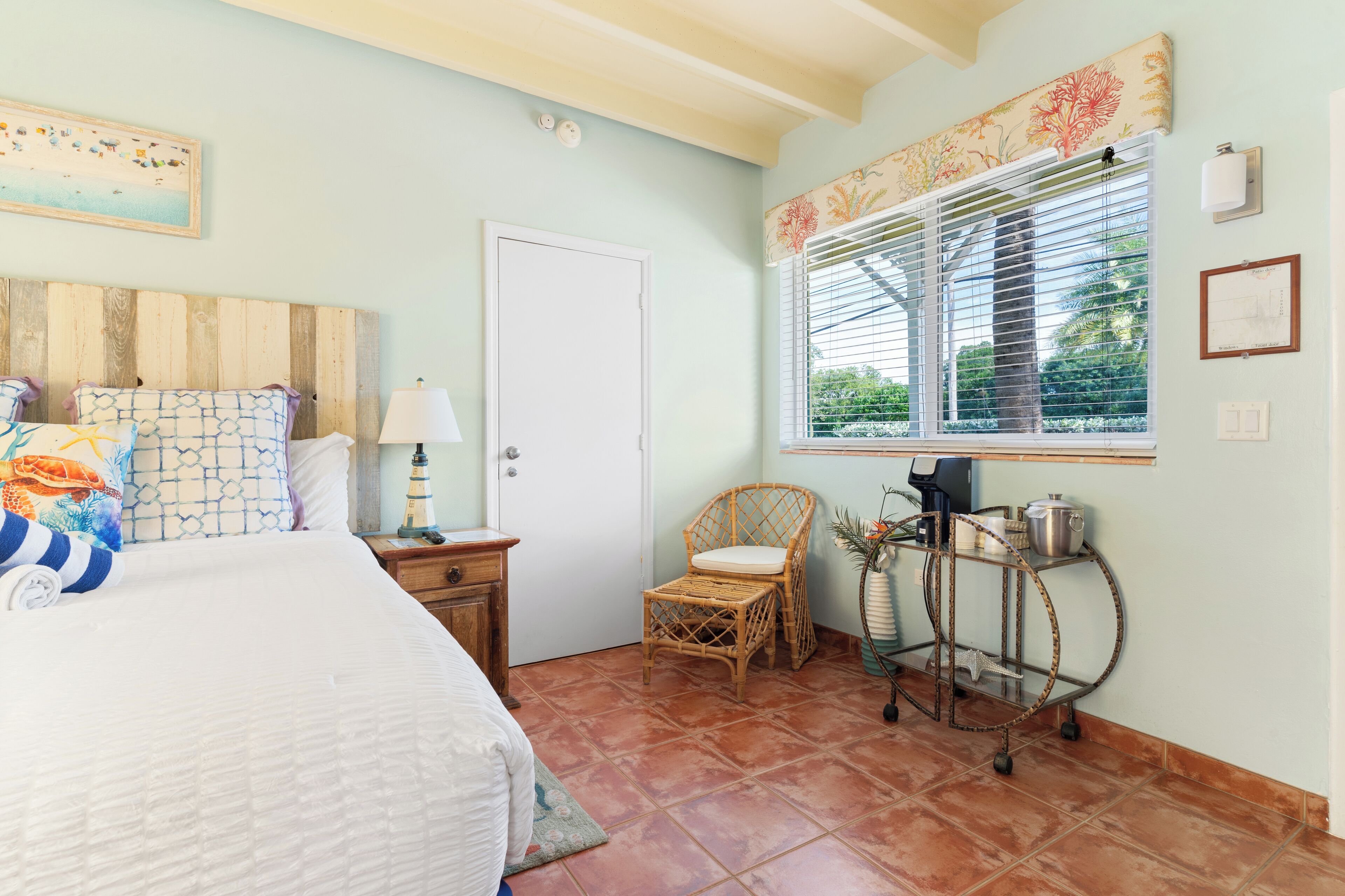 A bedroom with a bed, a nightstand, a chair, a window with blinds, and a wall-mounted shelf with kitchen items.