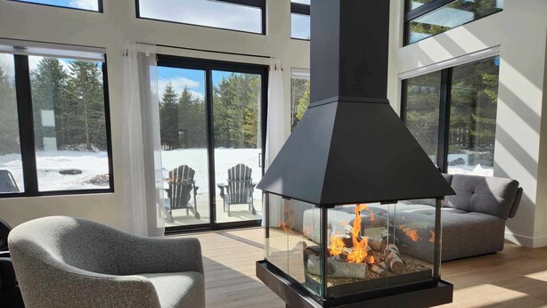 A modern living room with a glass fireplace, large windows, and a cozy seating area. - Ô 7e ciel spa and massage (weedon)