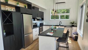A modern kitchen with dark cabinetry, a stainless steel refrigerator, a central island with bar stools, and a window providing natural light. - Ô 7e ciel spa and massage (weedon)