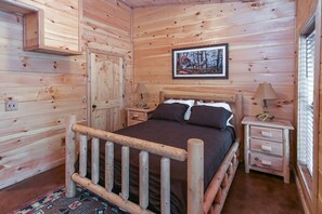 Habitación de cabaña de madera con cama, dos mesitas de noche, un tocador, una puerta y un cuadro enmarcado en la pared.
