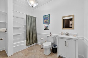 Une salle de bain avec des toilettes, un lavabo et un rideau de douche. On y trouve des serviettes sur une étagère, une poubelle et un tableau encadré au mur.