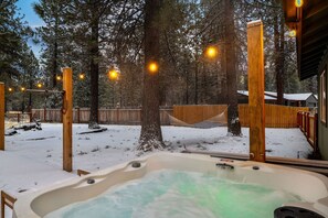 A hot tub in a snowy outdoor area with wooden structures and string lights. - Hot Tub, Fire Pit, Dog Friendly (Sisters)