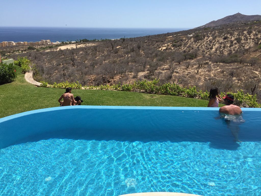 A swimming pool with clear blue water, surrounded by a grassy area and a view of a mountainous landscape.