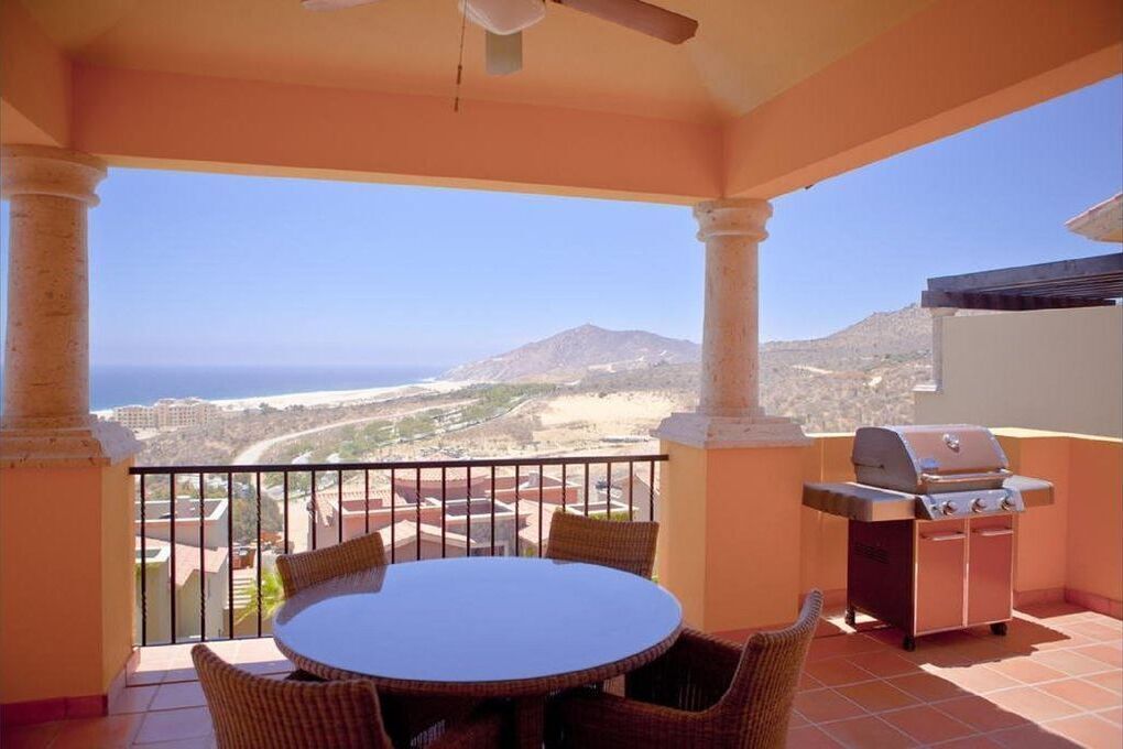 A balcony with a round table and chairs, a grill, and a view of a mountainous landscape.