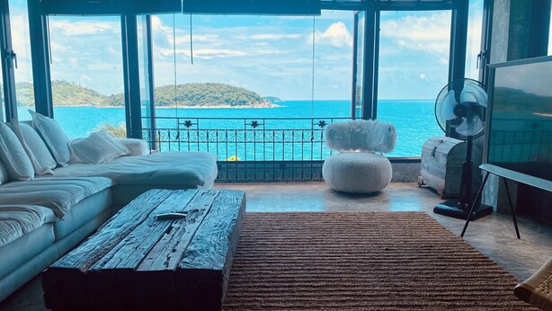 Un moderno salón con un amplio ventanal con vistas a la costa, un sofá blanco, una mesa de centro de madera y un otoman blanco y mullido.