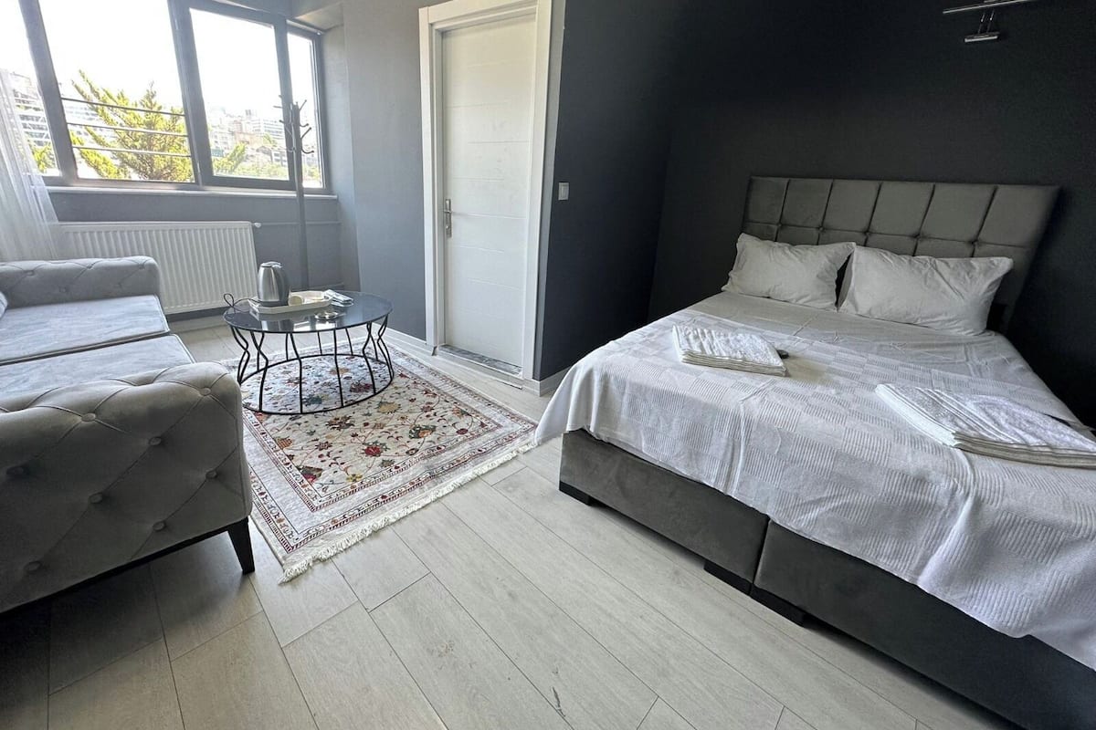 Ein modernes Schlafzimmer mit einem großen Bett, einer Couch, einem kleinen Tisch und Blick ins Freie durch ein Fenster.