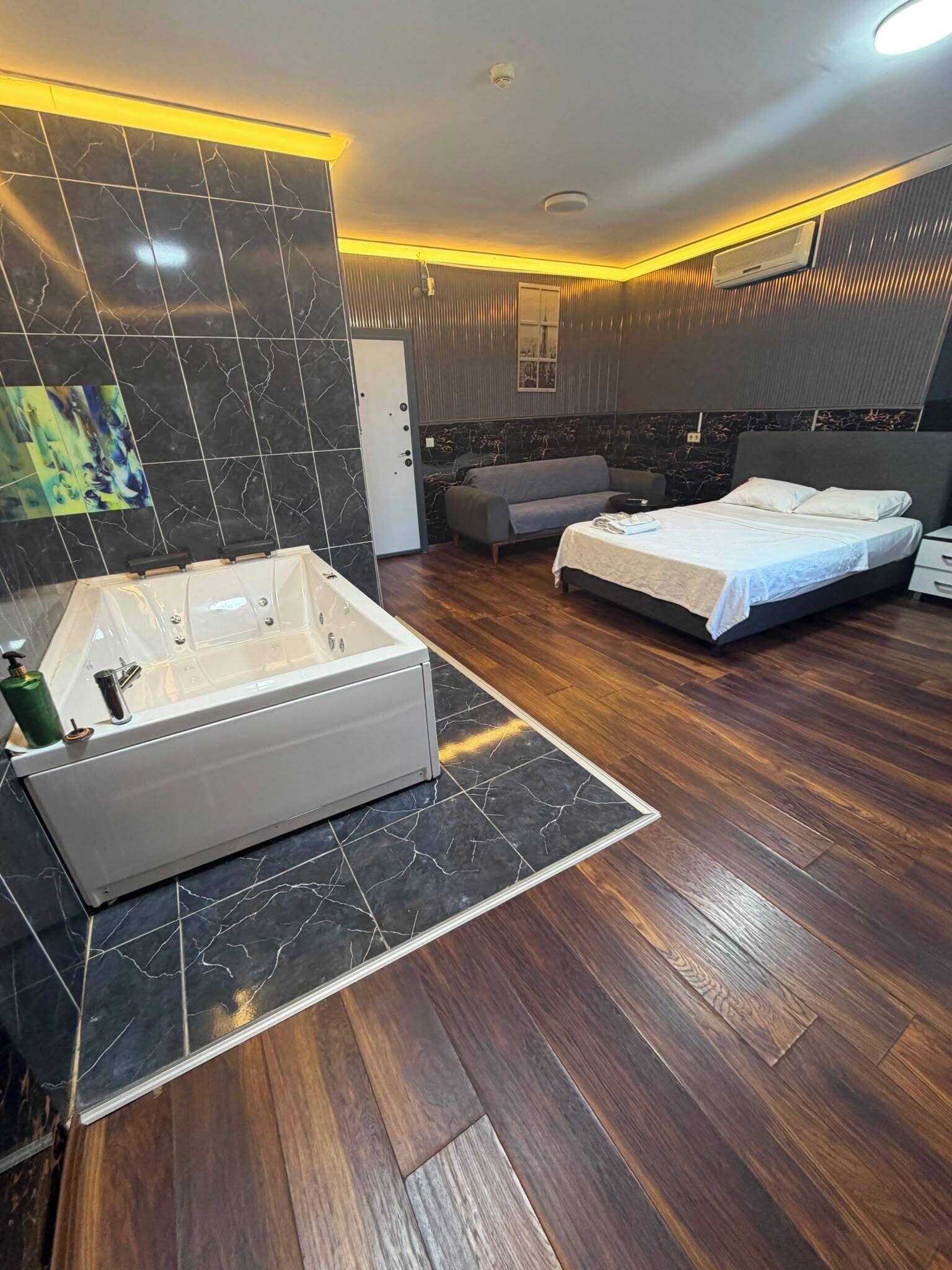 Une salle de bain moderne avec une grande baignoire, une douche et un meuble-lavabo. On y trouve un lit avec une tête de lit grise et une table de chevet. Le sol est en bois et les murs ont une finition sombre et texturée.