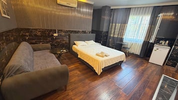 Habitación de hotel con cama, sofá, una mesita, kitchenette y ventana con cortinas.