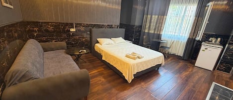 Habitación de hotel con cama, sofá, una mesita, kitchenette y ventana con cortinas.