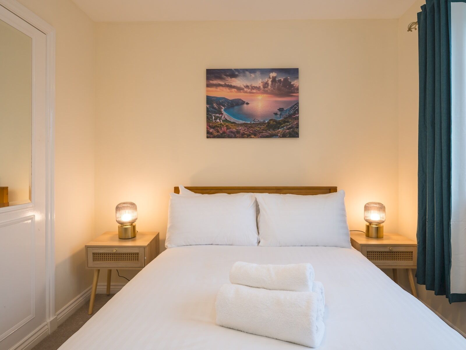 Ein Hotelzimmer mit einem großen Bett, zwei Nachttischen mit Lampen, einem Gemälde einer Küstenlandschaft und einem Fenster mit Vorhängen.