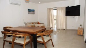 A modern living room with a wooden dining table and chairs, a sofa, a flat-screen TV, and a large window with curtains. - Sunset #105 Condominium - HD4 (Cabo San Lucas)