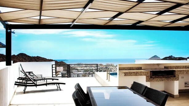 A rooftop terrace with a glass table and chairs, a grill, and a view of the coastline. - Sunset #105 Condominium - HD4 (Cabo San Lucas)