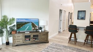 A living room with a flat-screen TV, a wooden cabinet, two bar stools, a ceiling fan, and a large window. - 2BR at Sandpiper Cove – Pool, Bay, Beachwalk (Englewood)