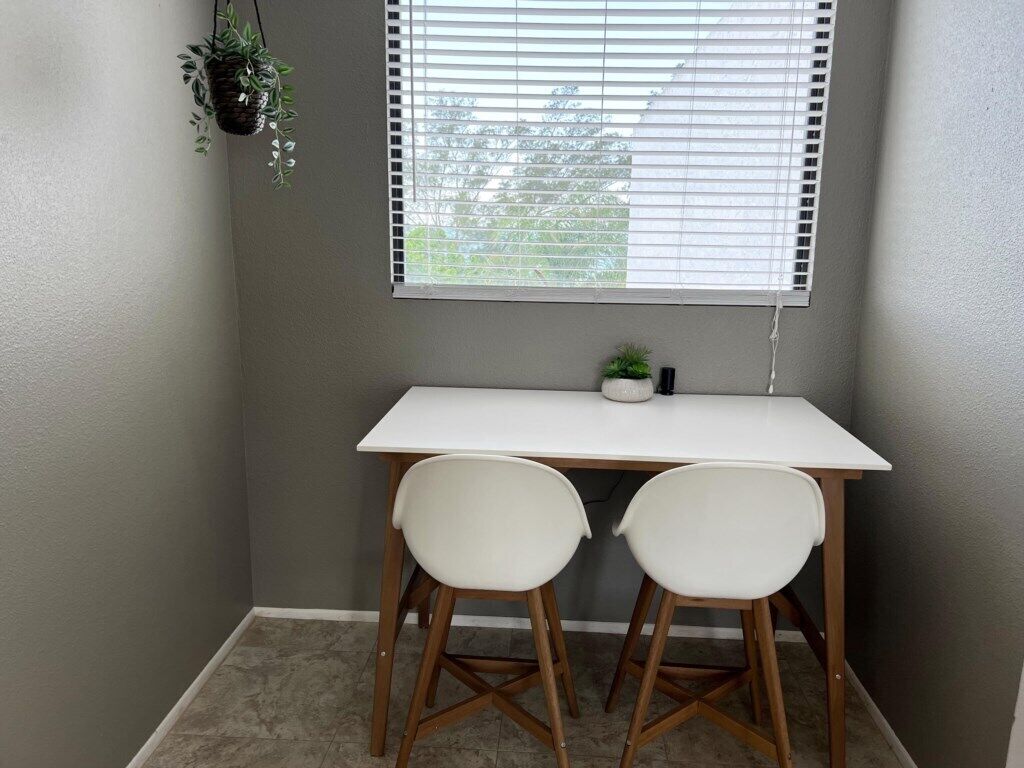 A small room with a white desk, two white chairs, a potted plant, and a window with blinds.