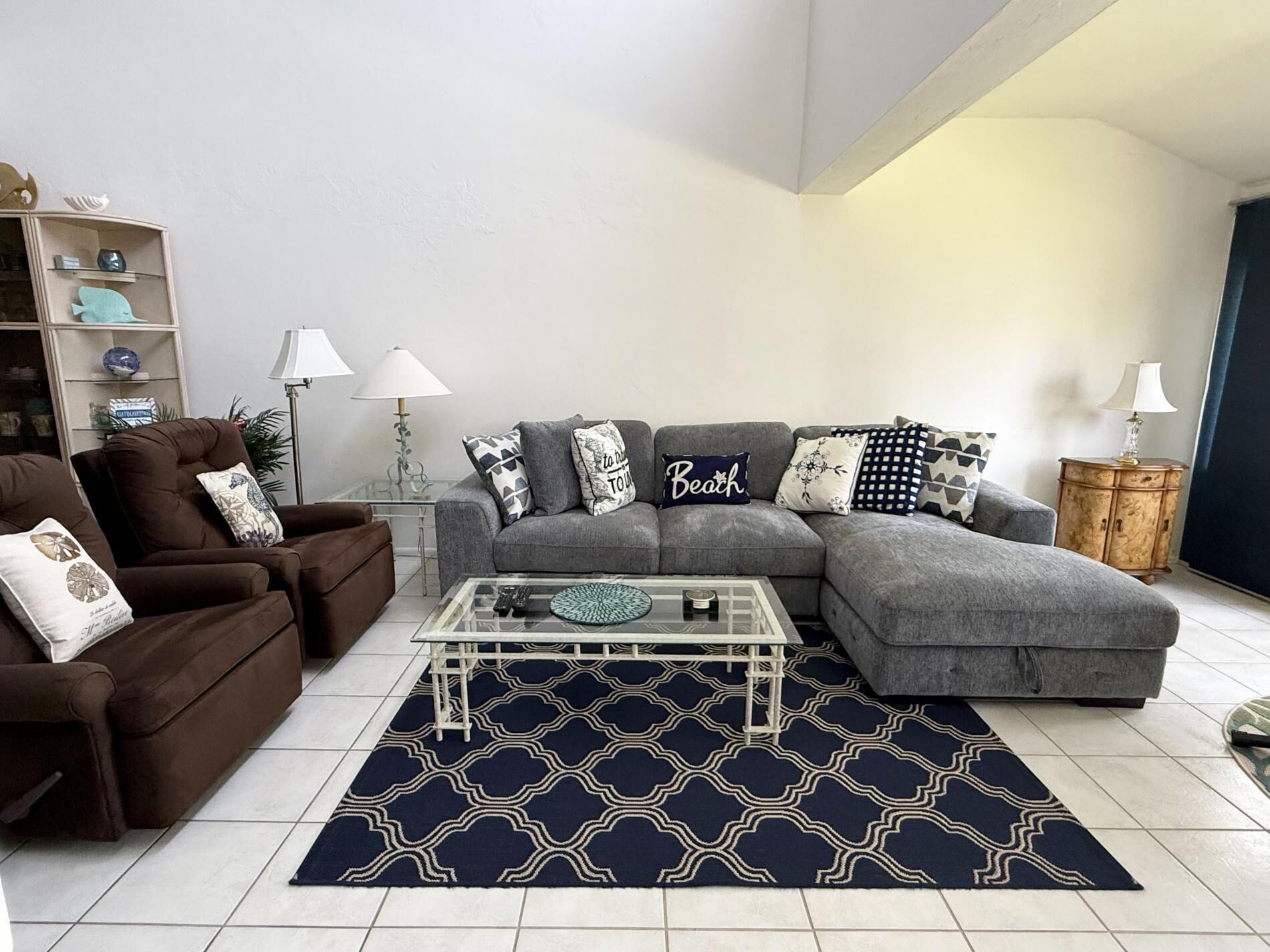 A living room with a grey sectional sofa, a brown armchair, a glass coffee table, and a patterned rug.