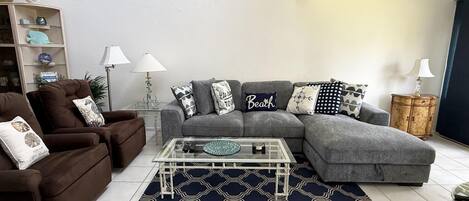 A living room with a grey sectional sofa, a brown armchair, a glass coffee table, and a patterned rug.