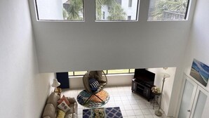 Una sala de estar con un sofá, una mesa de centro y un televisor. Tres ventanas proporcionan luz natural.