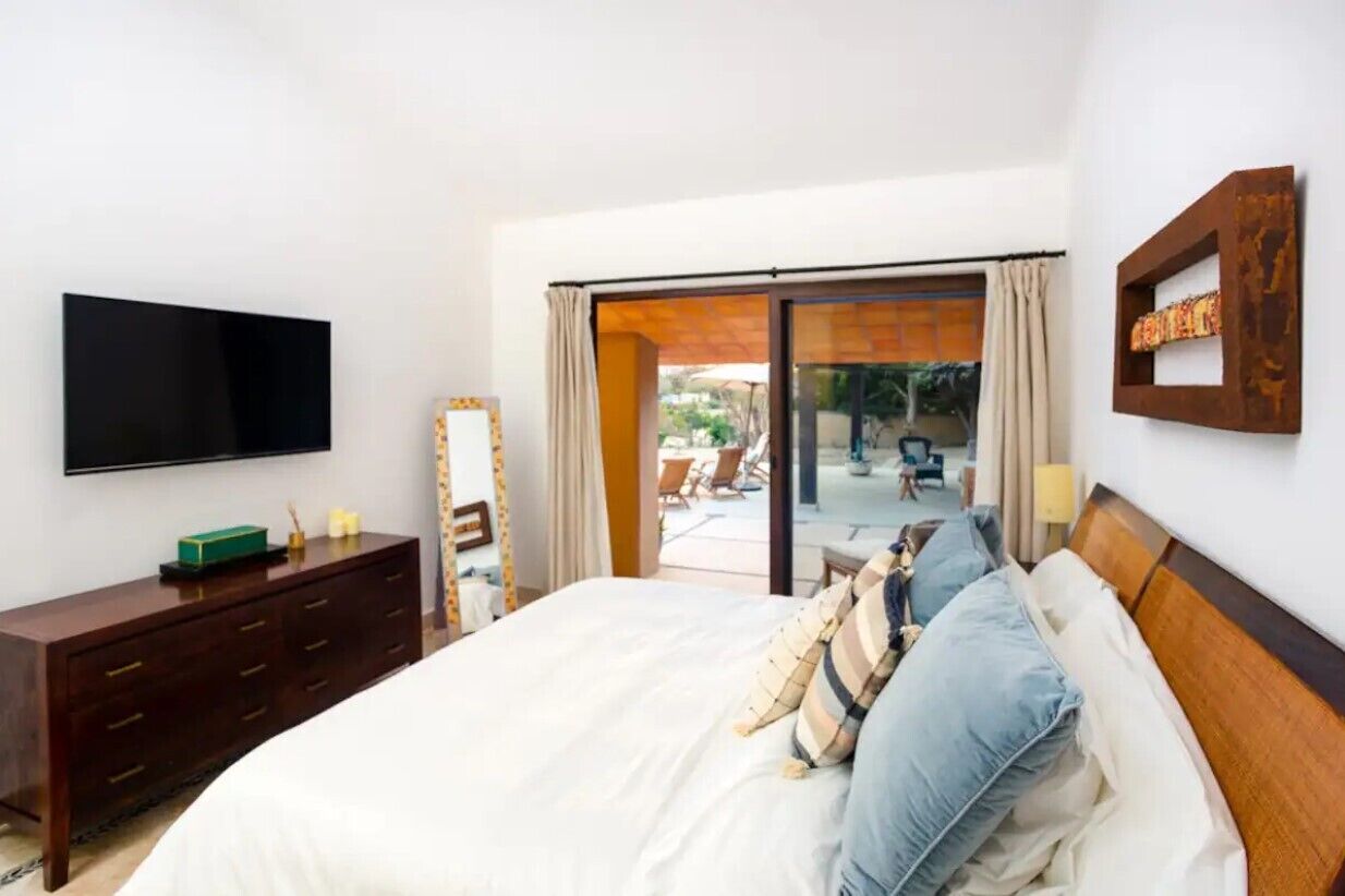 A bedroom with a large bed, a TV mounted on the wall, a wooden dresser, and a sliding glass door leading to an outdoor area with chairs and a table.