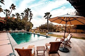 Uma piscina com espreguiçadeiras e um guarda-sol, rodeada por palmeiras e um jardim.