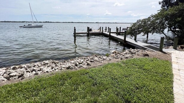 A marina with a dock extending into calm water, a sailboat anchored in the distance, and a grassy area with a paved walkway along the water's edge. - Lemon Bayview Villa - Heated Pool - Tennis - Dock (Englewood)