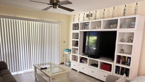 A living room with a flat-screen TV, white shelving with decorative items, a glass coffee table, and a ceiling fan. - Lemon Bayview Villa - Heated Pool - Tennis - Dock (Englewood)
