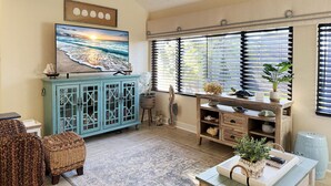 A living room with a large TV, a cabinet, wicker furniture, and a window with blinds. - Tamarind 3BR Loft – Beach Vibes & Bay Views (Englewood)
