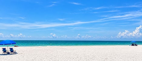 Una playa con arena blanca y un cielo azul despejado. Hay sombrillas y sillones azules.