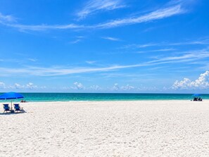 En strand med vit sand och en klarblå himmel. Här finns blåa parasoller och solstolar.
