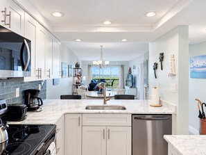 A modern kitchen with white cabinetry, a central island, and a view to the outside. - 2BR - Dolphins & Bay Views Just Steps to Beach (Bonita Springs)