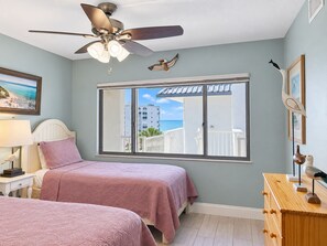 A bedroom with two beds, a ceiling fan, a window with a view of buildings and the sea, and a wooden dresser. - 2BR - Dolphins & Bay Views Just Steps to Beach (Bonita Springs)