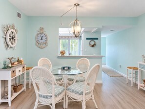 A dining area with a round glass table surrounded by wicker chairs, a wall clock, and a chandelier.