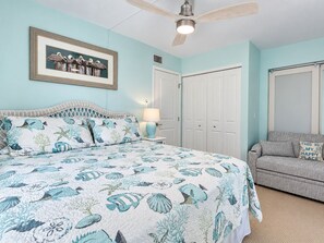 A bedroom with a bed, a ceiling fan, a framed picture, a nightstand, a lamp, a couch, and a door.